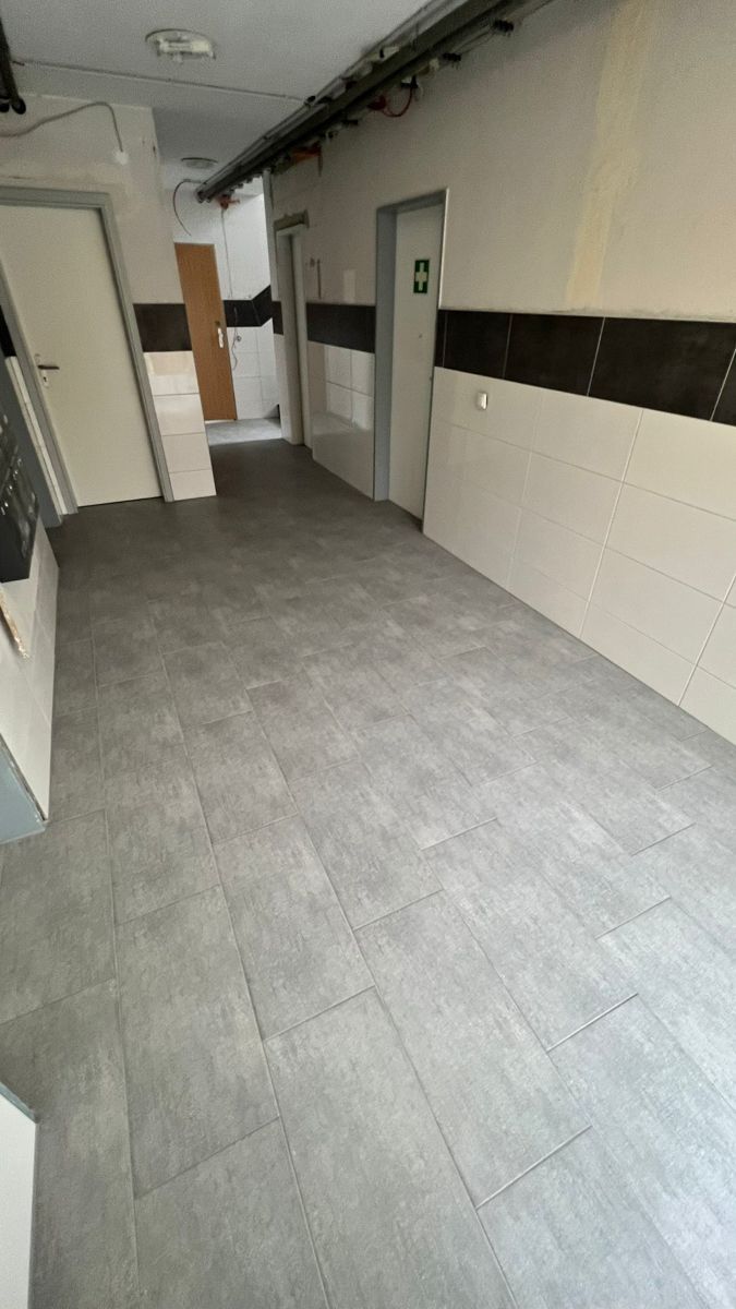 Leerer Flur mit grauem Fliesenboden, weißen Wänden und schwarzen Zierleisten.