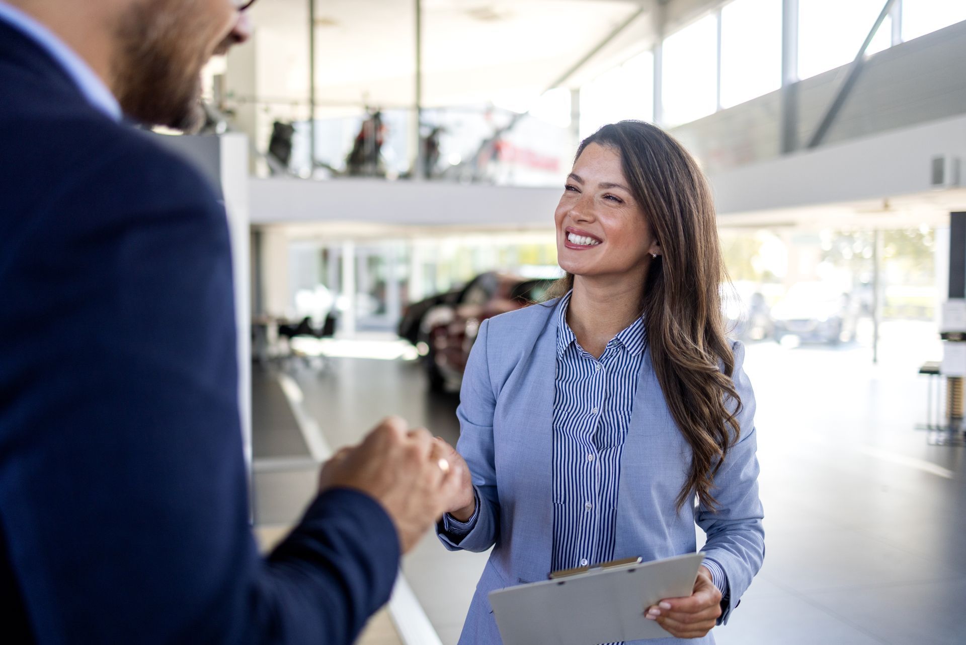 Une femme en blazer bleu tape dans la main d'un homme dans une concession automobile, souriante et tenant un bloc-notes.