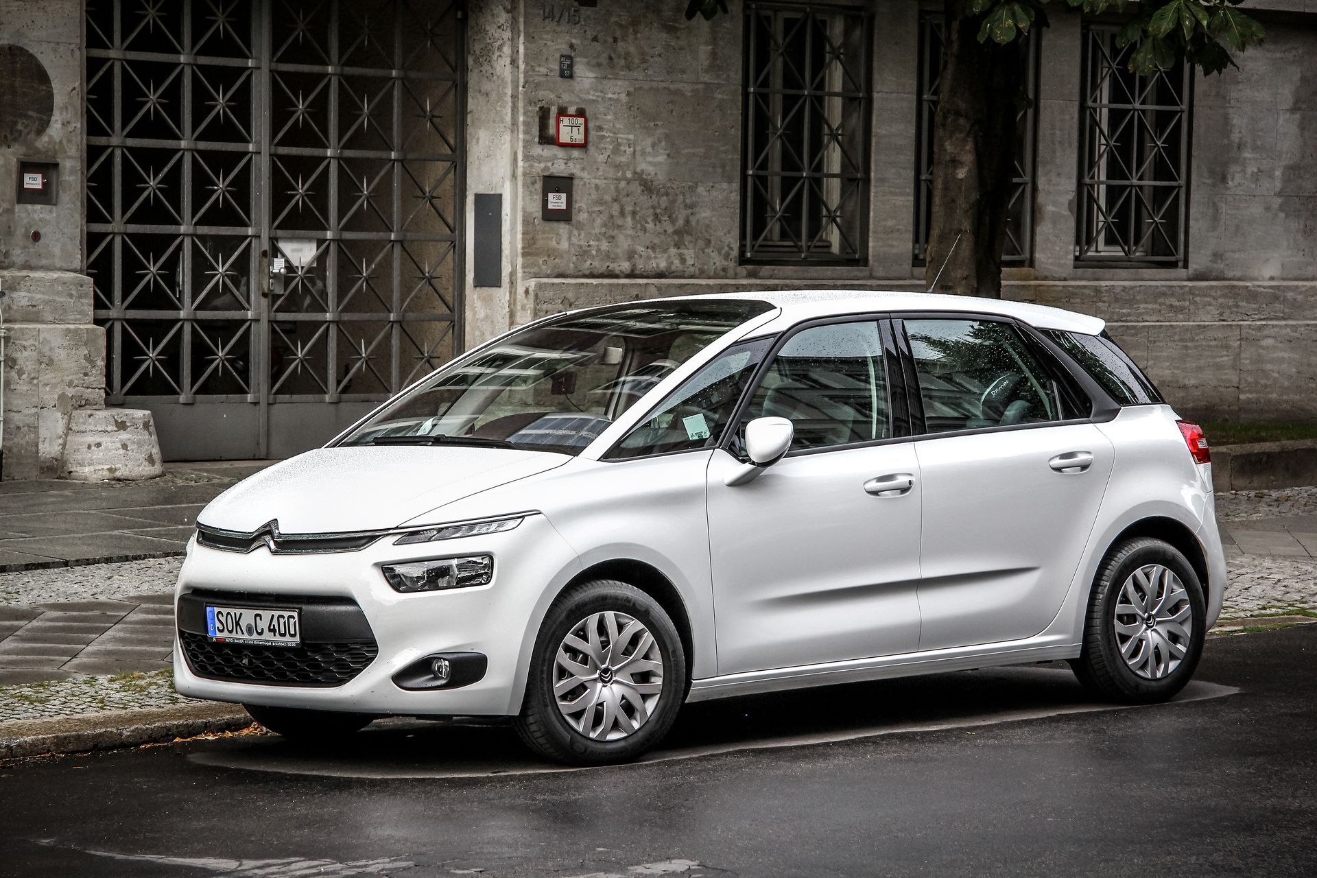 Citroën C4 Picasso blanche garée dans une rue de la ville, devant un immeuble aux portes ornées.
