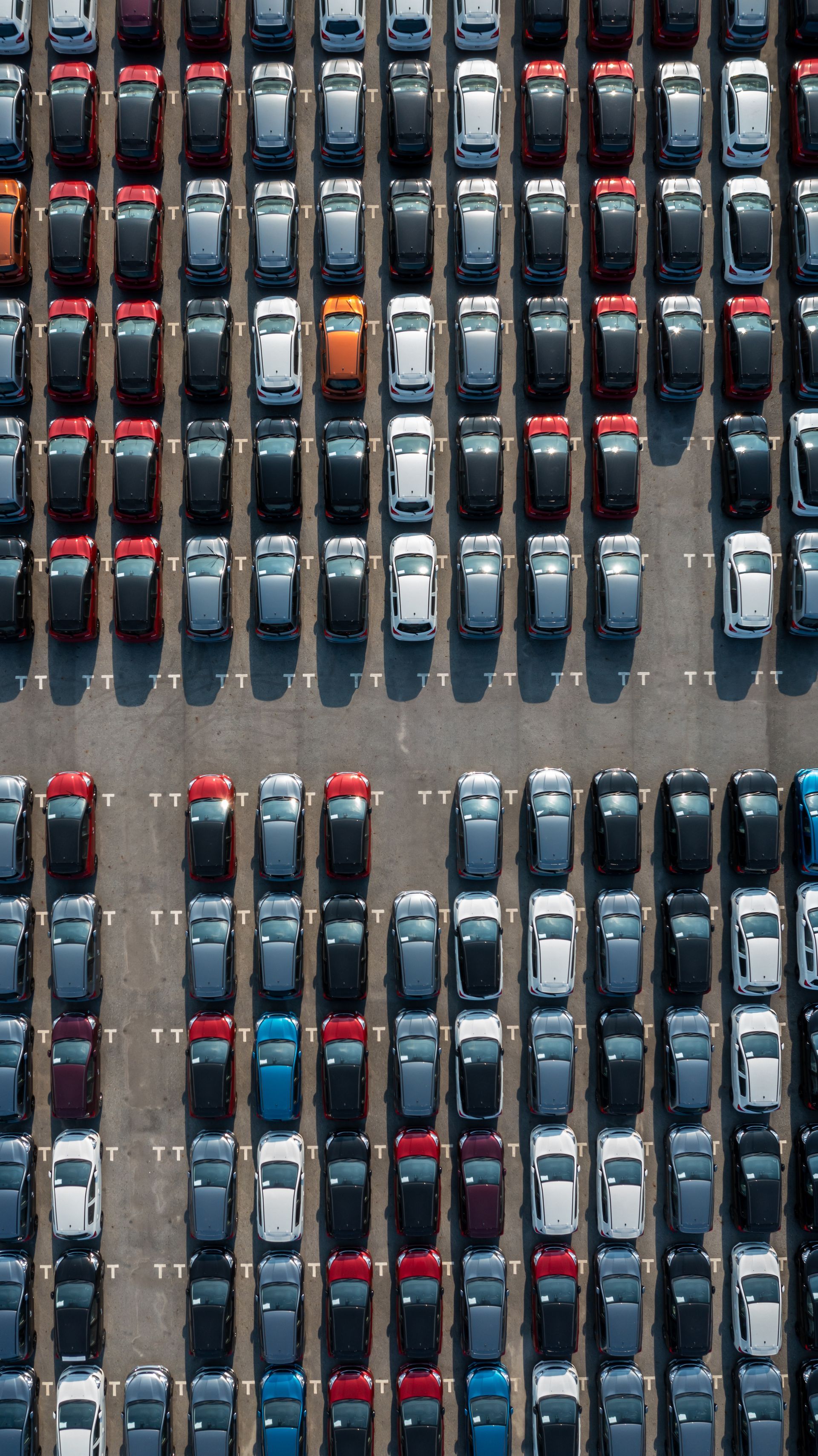 Vue aérienne d'un parking rempli de rangées de voitures neuves de différentes couleurs : grises, noires et rouges.