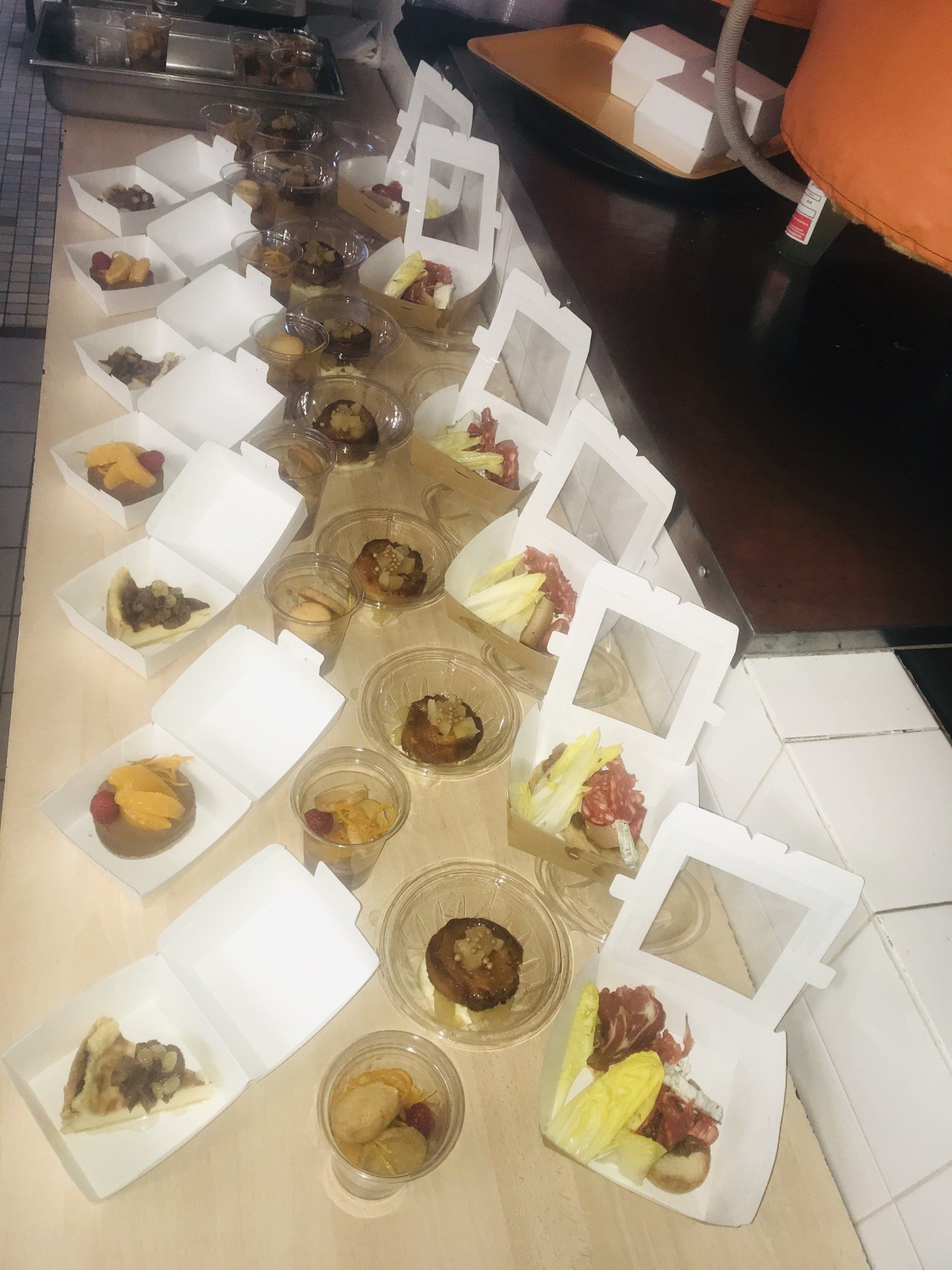 Présentation de plats traiteur avec de petites boîtes blanches et des bols transparents contenant divers mets sur une table beige.