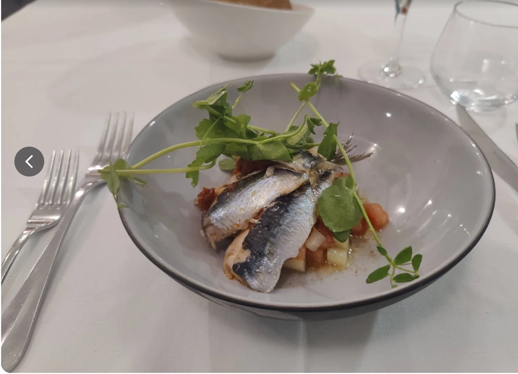 Sardines servies dans un bol gris avec de la verdure, accompagnées de couverts et de verres sur une nappe blanche.