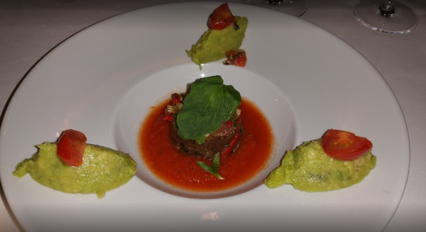 Assiette élégante avec un plat central de sauce rouge et de garniture verte, entouré d'éléments à base d'avocat et de demi-tomates.