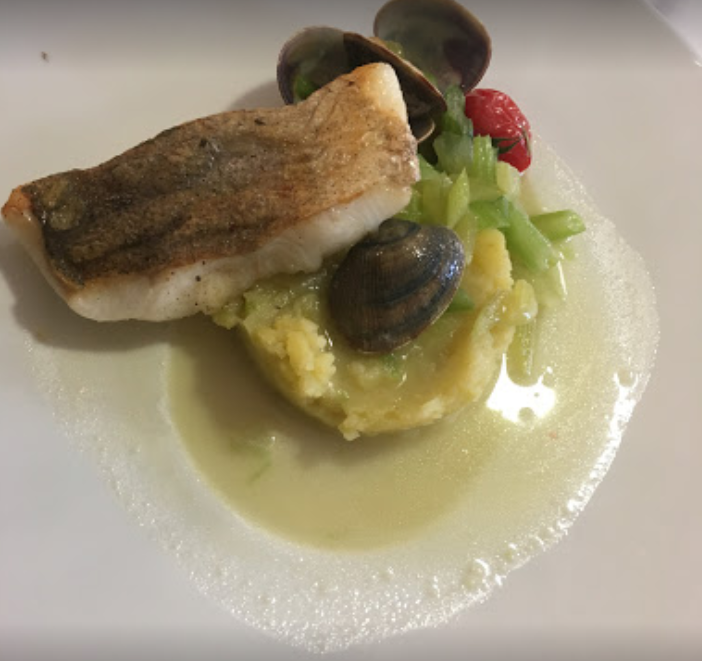 Poisson poêlé sur un lit de purée de pommes de terre, avec des palourdes et de la sauce, servi dans une assiette blanche.