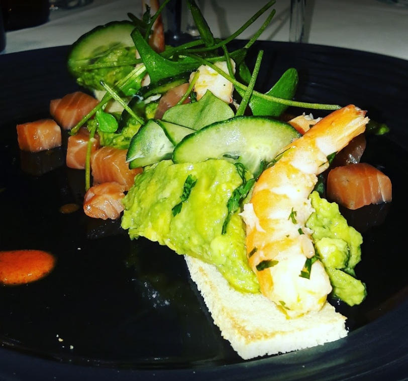 Salade de fruits de mer sur assiette noire : cubes de saumon, crevettes, avocat, concombre, salade verte et toast.
