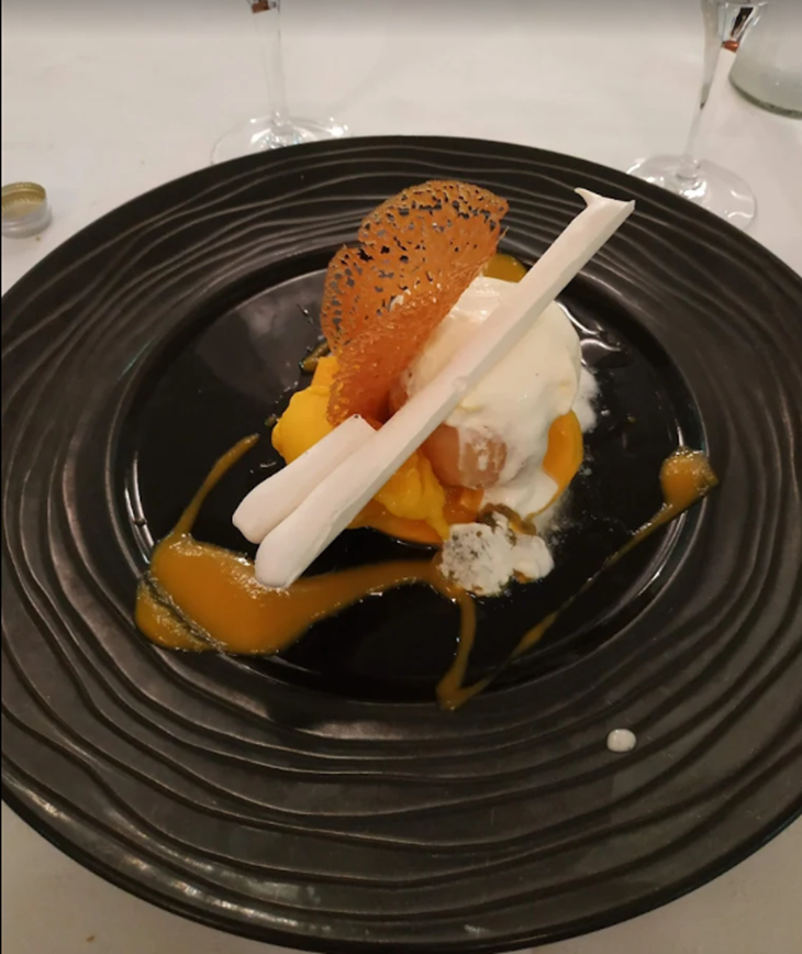 Dessert servi sur une assiette noire : purée de mangue, bâtonnets de meringue, glace et tuile décorative.
