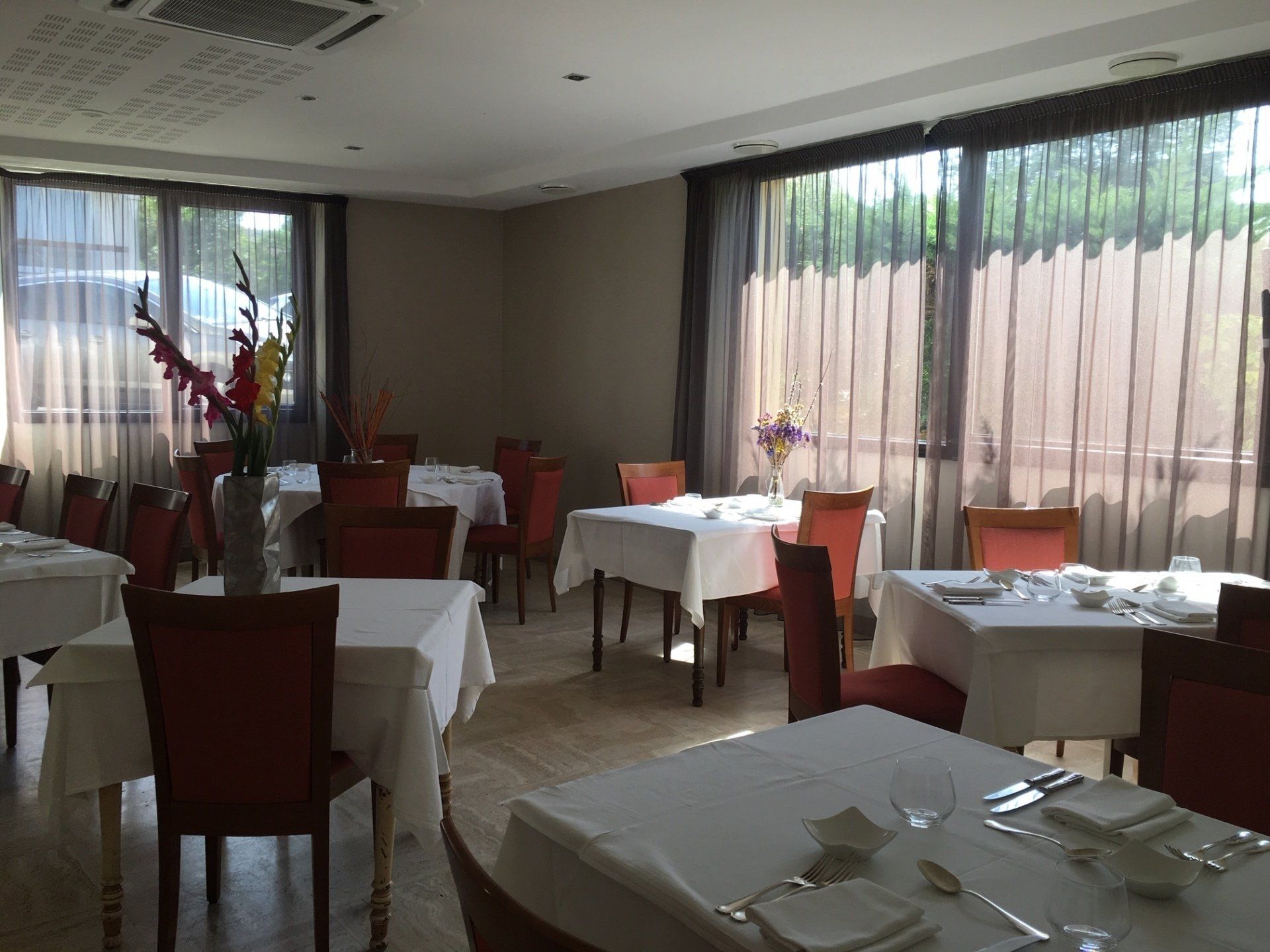 Salle à manger du restaurant avec tables dressées pour les clients, fenêtres avec voilages et chaises orange.