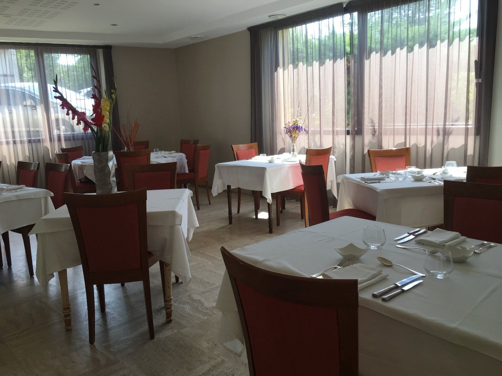 Salle à manger du restaurant avec nappes blanches, chaises rouges et grandes fenêtres aux rideaux transparents.