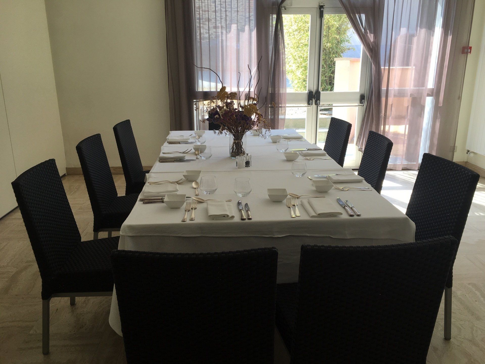 Table à manger dressée pour un repas avec huit chaises noires, nappe blanche et fenêtre en arrière-plan.