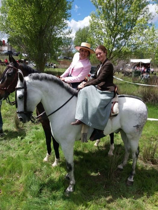 Dos personas montando caballos blancos en un parque cubierto de hierba; una de ellas llevaba una camisa rosa y un sombrero de paja.