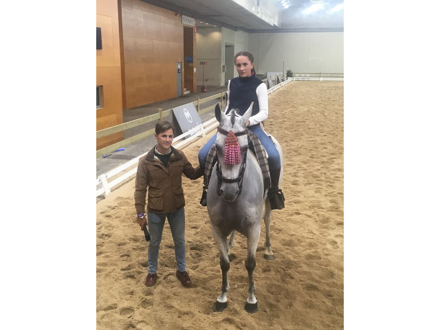 Una persona monta un caballo gris, guiada por otra persona, en una pista de equitación cubierta.