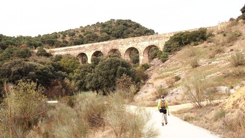 Acueducto de piedra en la ladera de una colina, sobre un sendero de tierra, con un excursionista solitario caminando debajo.