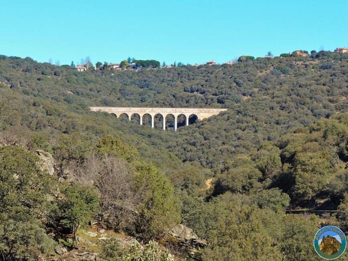 Viaducto de arcos de piedra que cruza un valle verde y boscoso bajo un cielo azul claro.