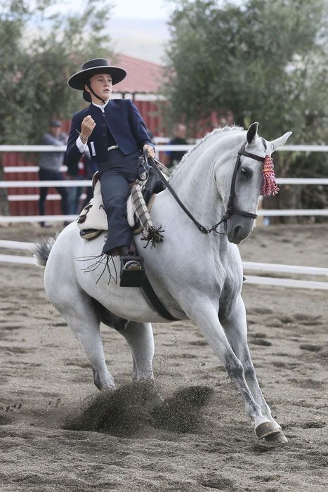 Una vaquera montando un caballo blanco en una arena, sosteniendo una rienda adornada con una cinta durante un espectáculo.