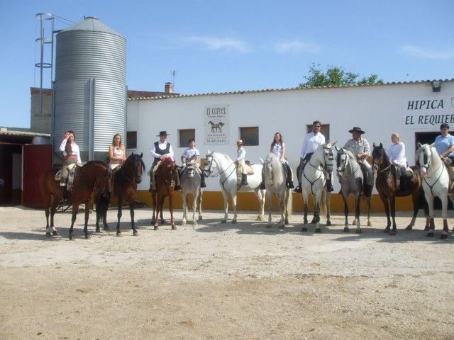 Varias personas a caballo formaban una fila frente a un edificio blanco con un silo en un patio soleado.