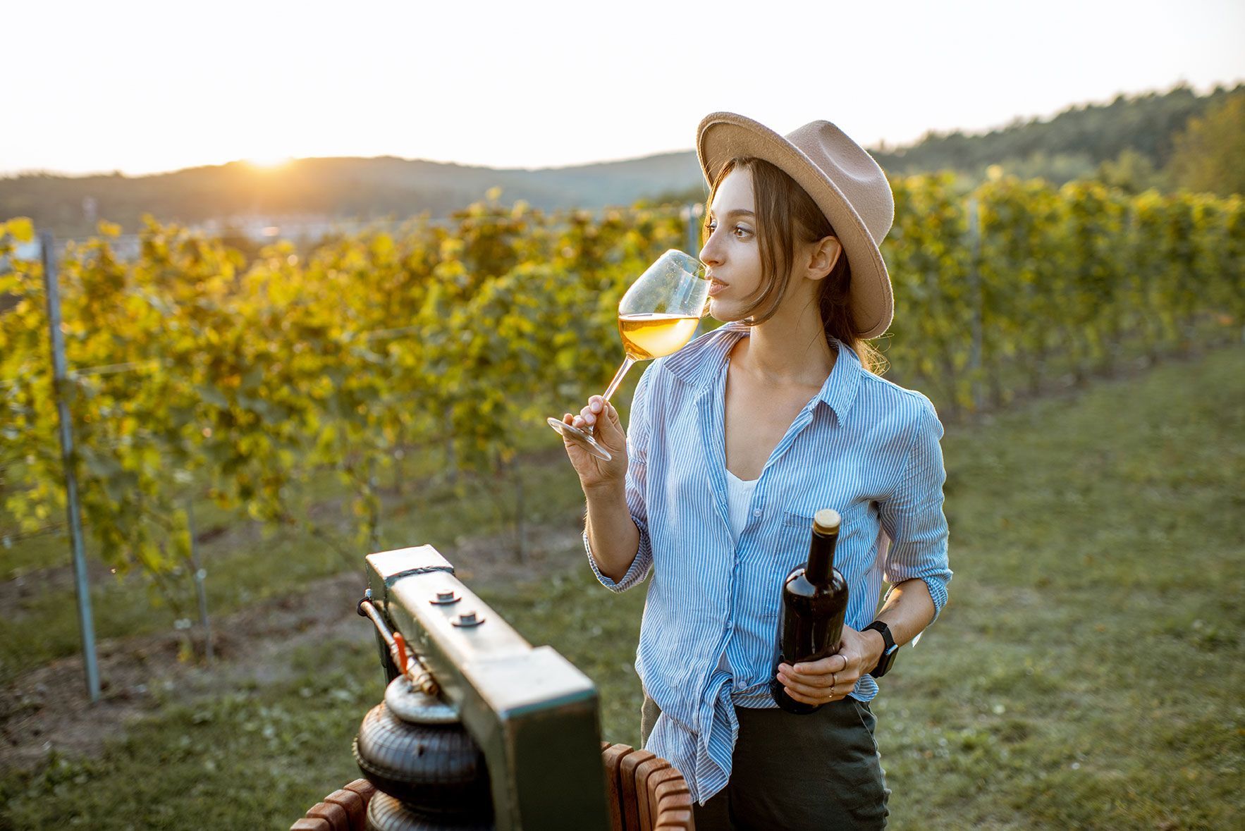 A woman holding a bottle of wine and a glass of wine in vineyard at sunset
