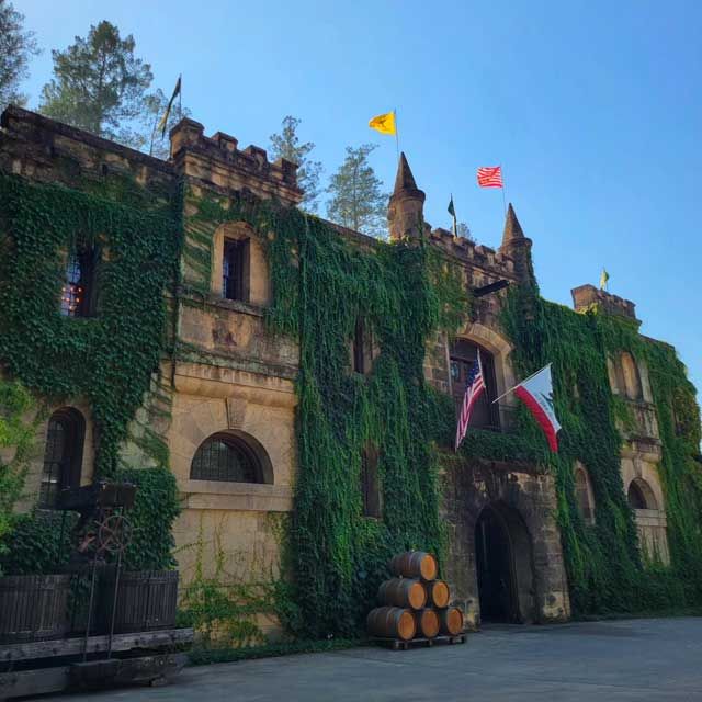 Chateau Montelena, a stone winery with ivy-clad Gothic architecture, flying flags, and wine barrels near the entrance, set against a blue sky