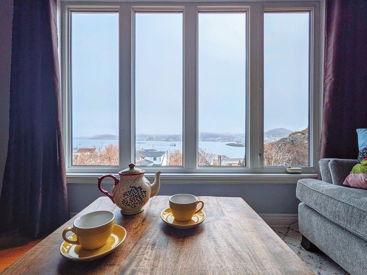 A coffee table with two cups and saucers and a teapot on it in front of a window with an ocean view