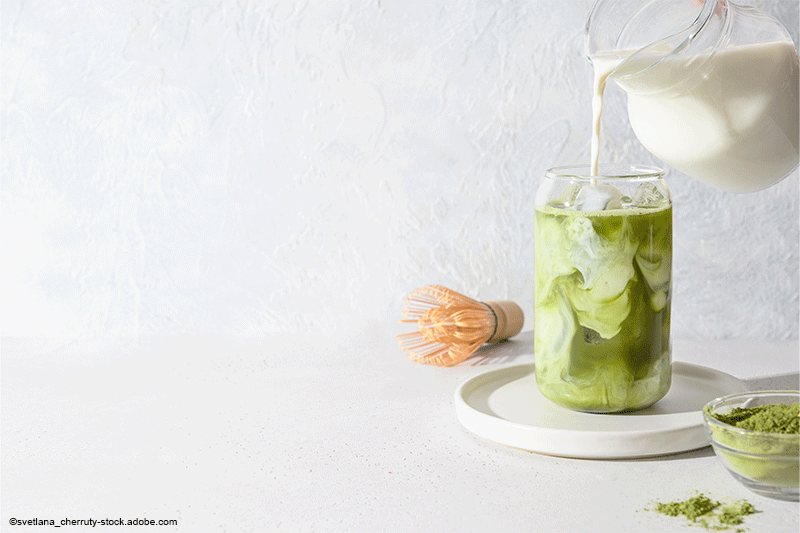 Milch wird in ein hohes Glas Matcha gegossen, daneben Schneebesen und Matcha-Pulver auf weißem Hintergrund.