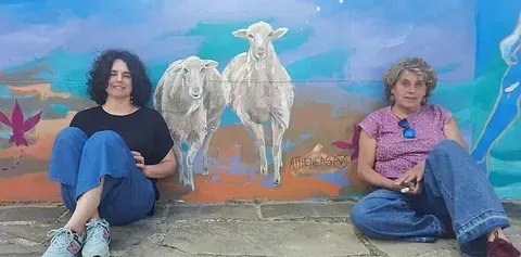 Dos mujeres están sentadas en el suelo frente a un mural de ovejas.