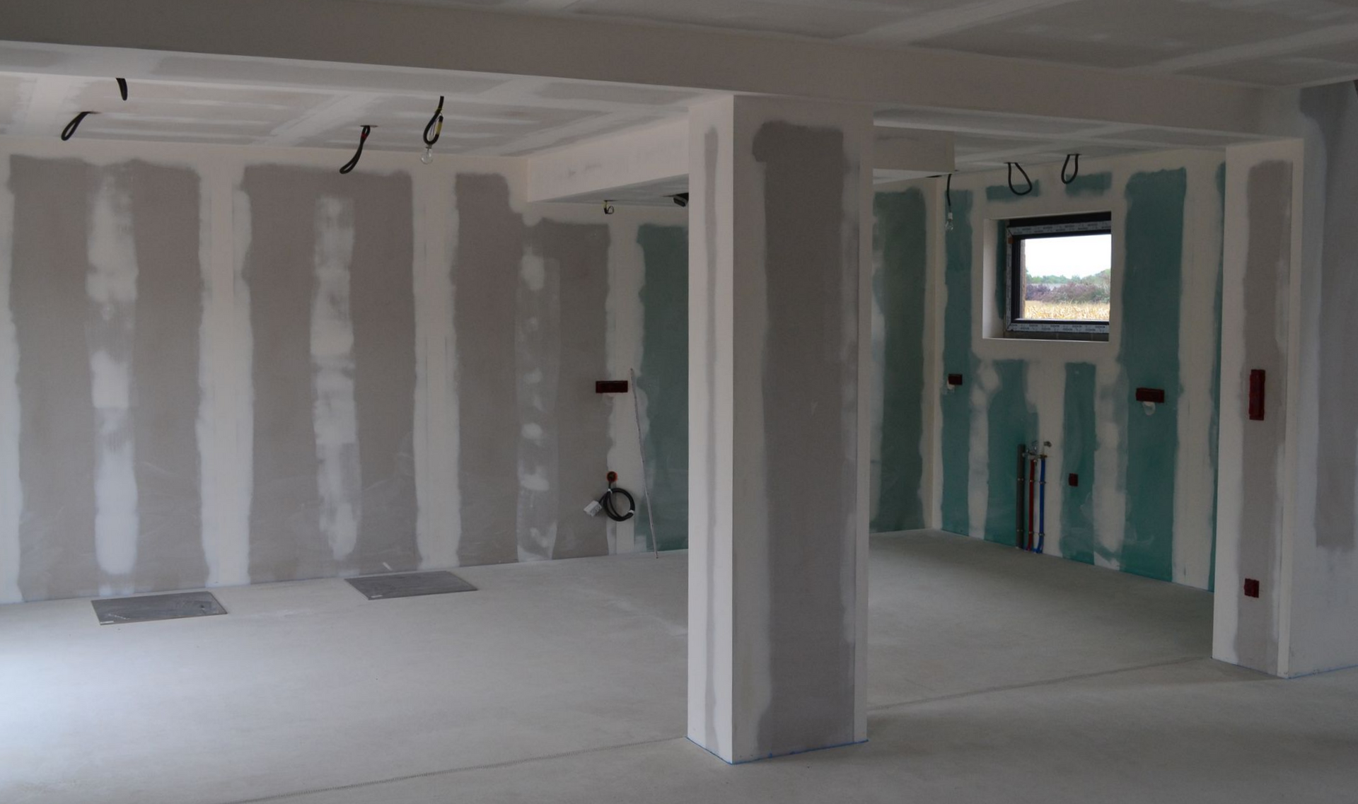 Intérieur d'une pièce en construction, avec des plaques de plâtre installées aux murs et au plafond, près d'une fenêtre.