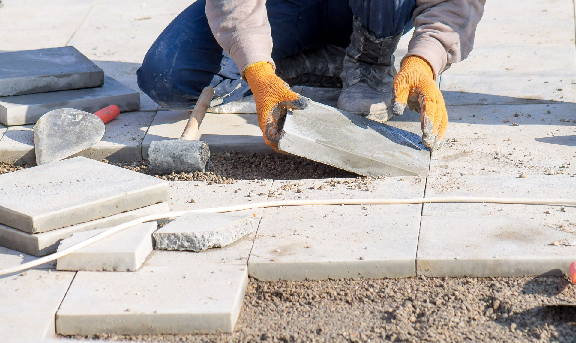 Personne posant des pavés gris à l'extérieur, portant des gants orange et un jean.