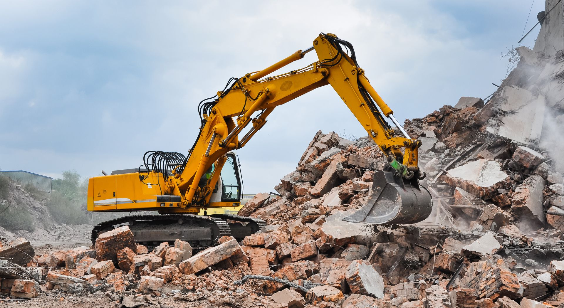 Une pelleteuse jaune démolit un bâtiment, entourée de décombres sous un ciel nuageux.
