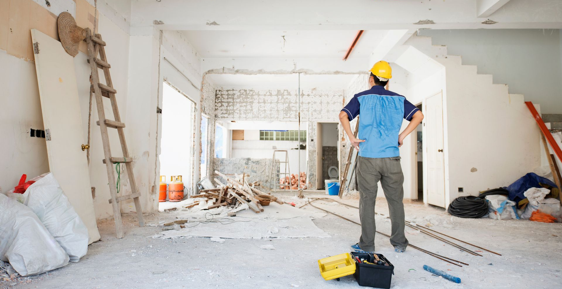 Un ouvrier du bâtiment, casque de chantier sur la tête, inspecte une pièce en rénovation ; des débris et des matériaux sont éparpillés.