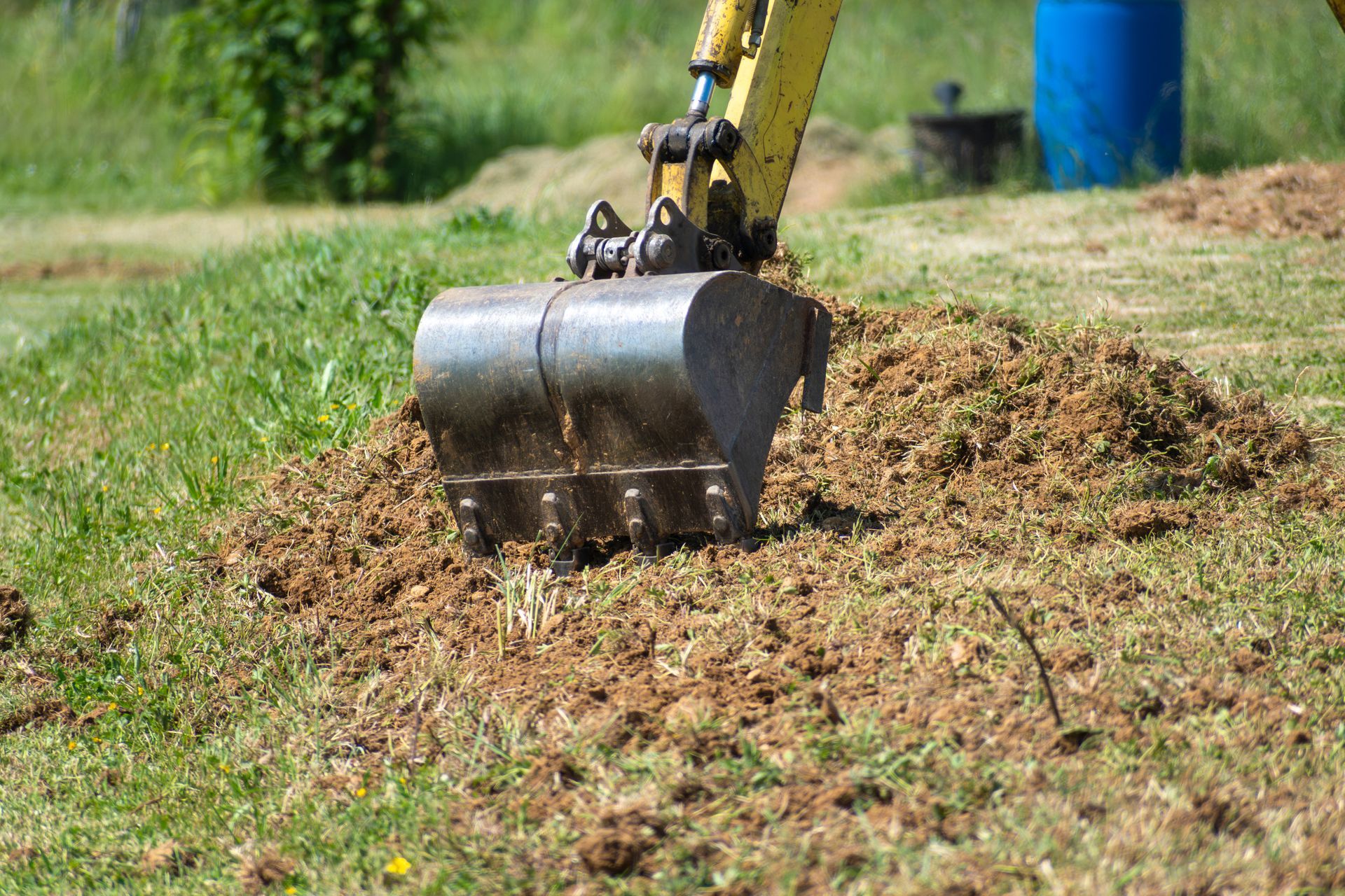 Godet d'une excavatrice creusant le sol dans une zone herbeuse ; baril bleu visible en arrière-plan.