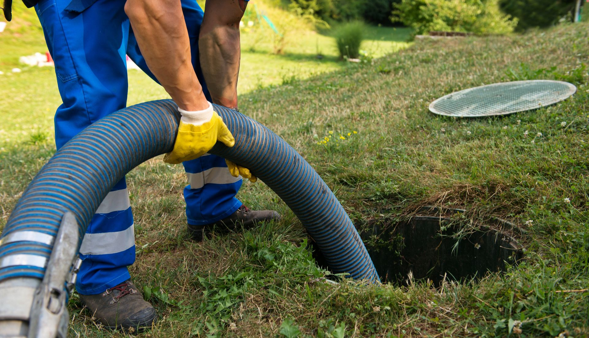 Un homme en salopette bleue vide une fosse septique avec un gros tuyau d'arrosage dans une cour herbeuse.