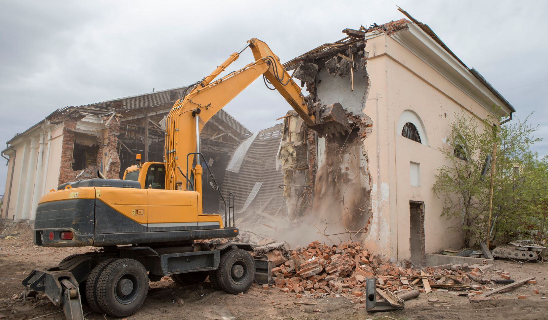 Une pelleteuse jaune démolit un bâtiment beige, soulevant poussière et débris.