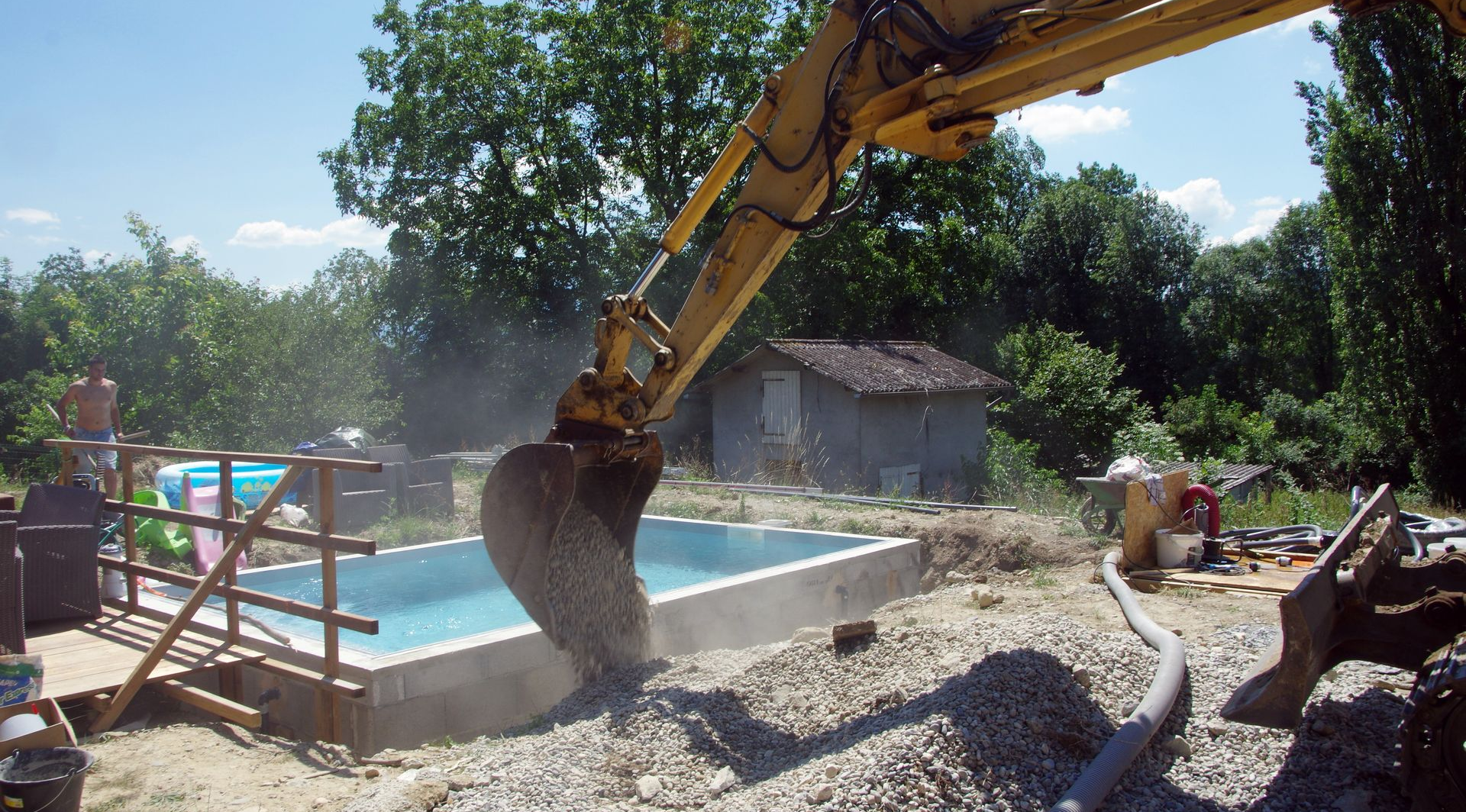 Une pelleteuse creuse près d'une piscine rectangulaire partiellement construite, dans un cadre extérieur ensoleillé.