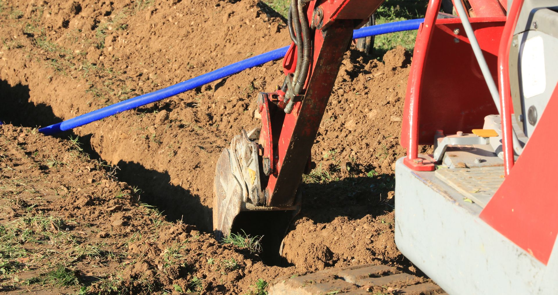 Une pelleteuse rouge creuse une tranchée et pose un tuyau bleu dans une zone herbeuse.