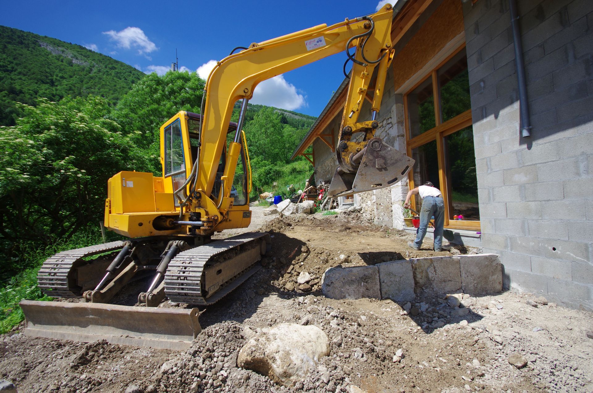 Une pelleteuse jaune démolit le mur d'un bâtiment, à côté d'une personne, avec des montagnes en arrière-plan.