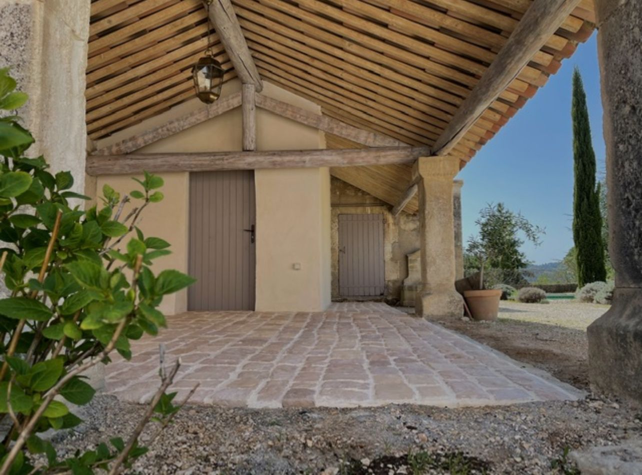 Véranda couverte avec sol en briques, poutres en bois, murs beiges et deux portes grises ; cadre extérieur avec verdure et ciel bleu.