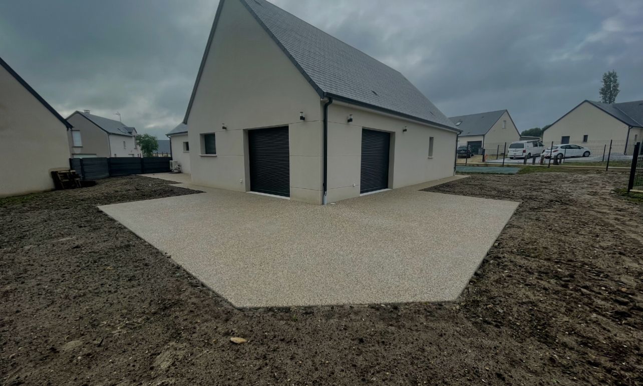Allée de gravier devant une maison claire au toit gris foncé et dotée de deux portes de garage. Ciel couvert.