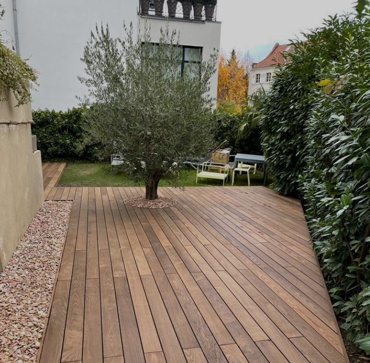 Terrasse en bois avec un olivier dans un jardin, bordée de verdure et d'une bordure de gravier.