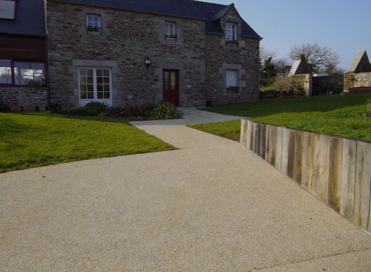 Maison en pierre avec allée de gravier et mur de soutènement en bois, entourée de pelouse verte.