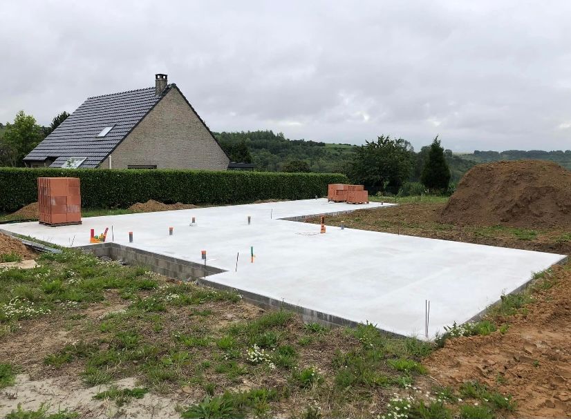 Fondations en béton d'une maison, avec une maison en arrière-plan. Des briques sont visibles sur les fondations. Journée nuageuse.
