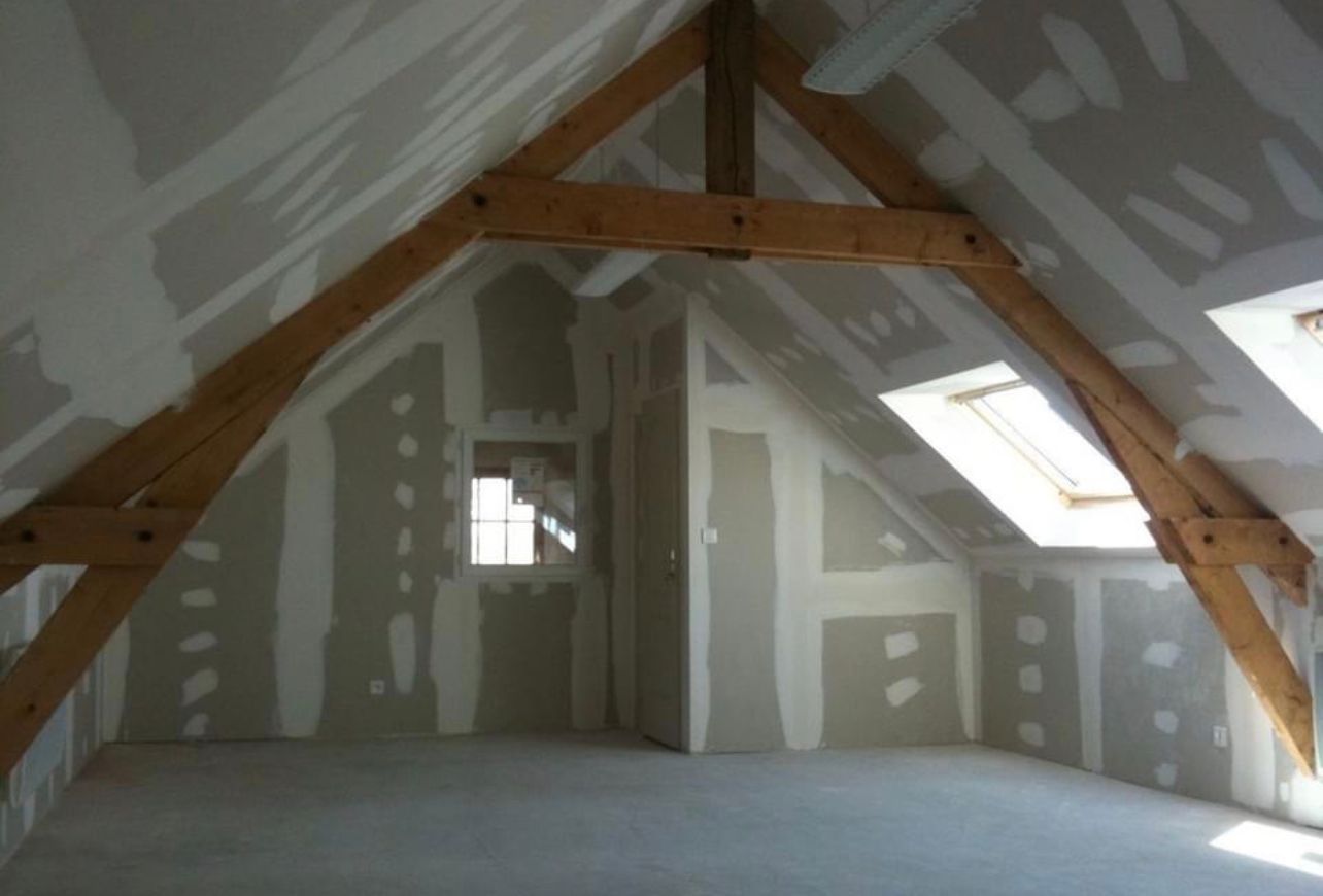 Vue intérieure d'un grenier avec poutres apparentes, murs en plaques de plâtre et une fenêtre.
