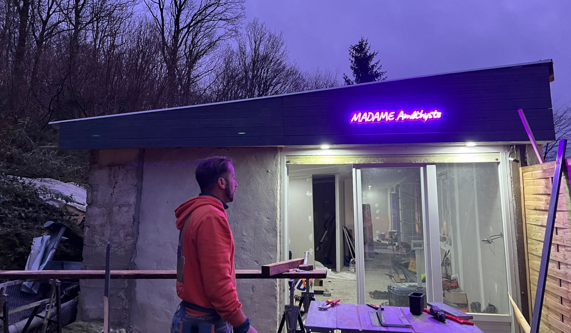 Un homme en chemise orange se tient devant un petit bâtiment orné d'une enseigne lumineuse violette indiquant MADAME Audigito.