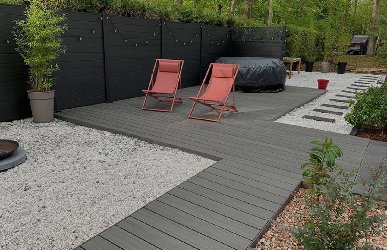 Terrasse moderne dans le jardin avec terrasse en composite gris, deux chaises orange, un jacuzzi et un chemin de gravier.