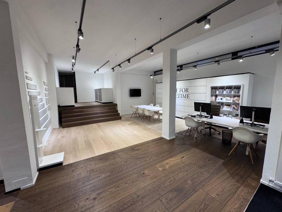 Interno di un moderno ufficio con pavimenti in legno, pareti bianche e illuminazione a binario. Sono presenti scrivanie e sedie.