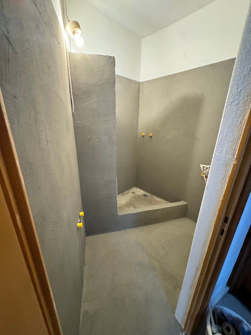 Piccolo bagno in costruzione con pareti in intonaco grigio e zona doccia triangolare.