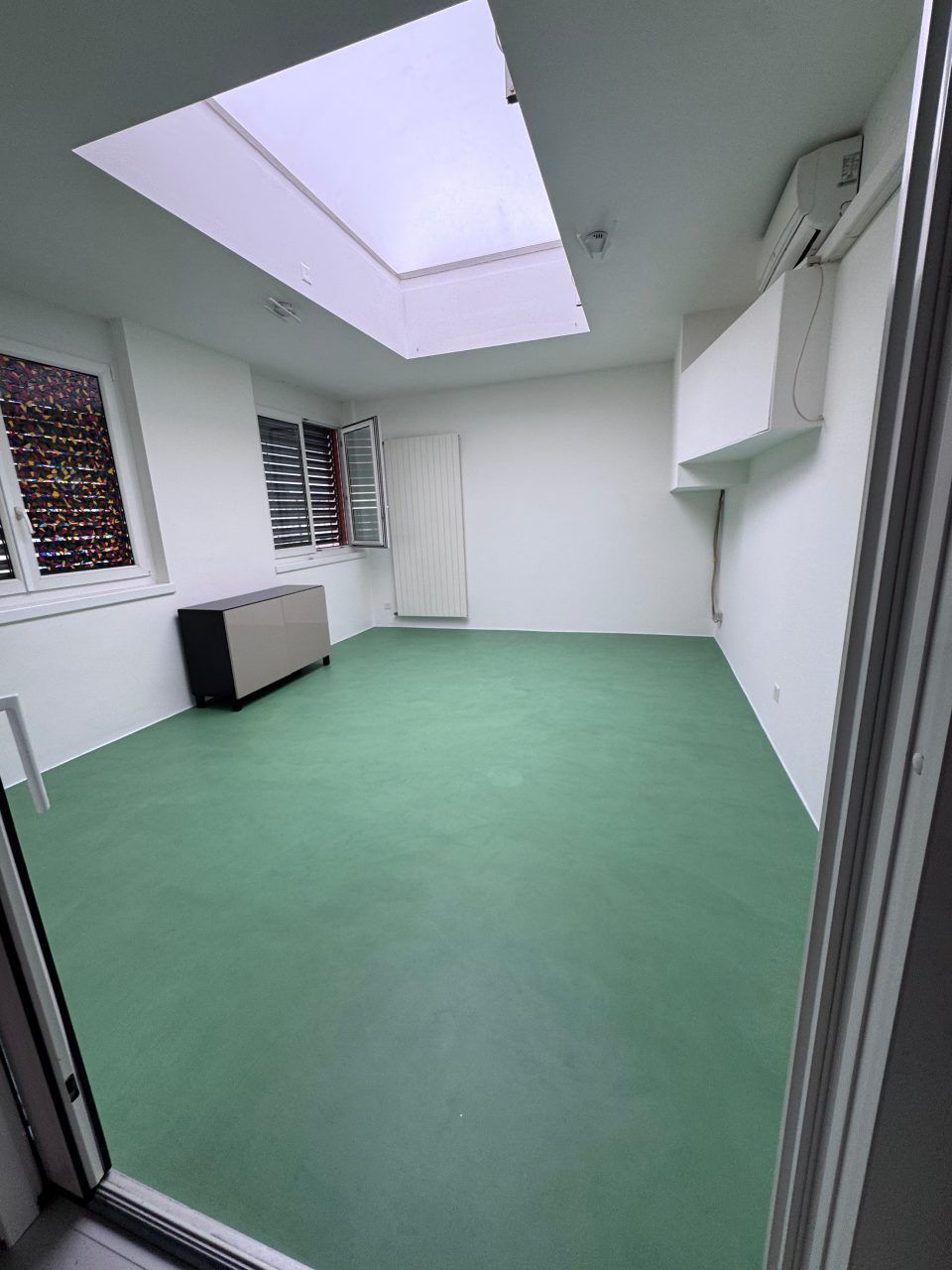 Stanza vuota con pavimento verde, pareti bianche, lucernario e piccola finestra.
