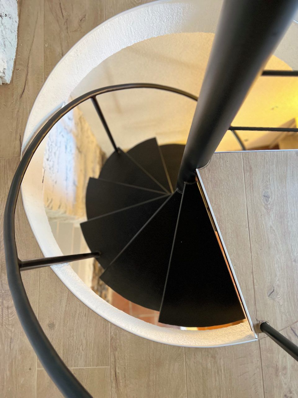 Vista dall'alto di una scala a chiocciola nera. La scala è circondata da una parete circolare bianca e da un pavimento in legno chiaro.