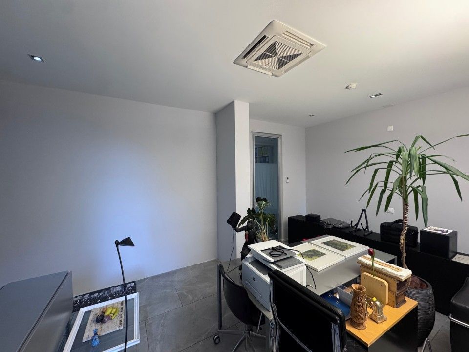 Ufficio con pareti bianche, pavimento grigio, aria condizionata. Nella stanza sono presenti piante e una scrivania con schermi per computer e altri materiali.