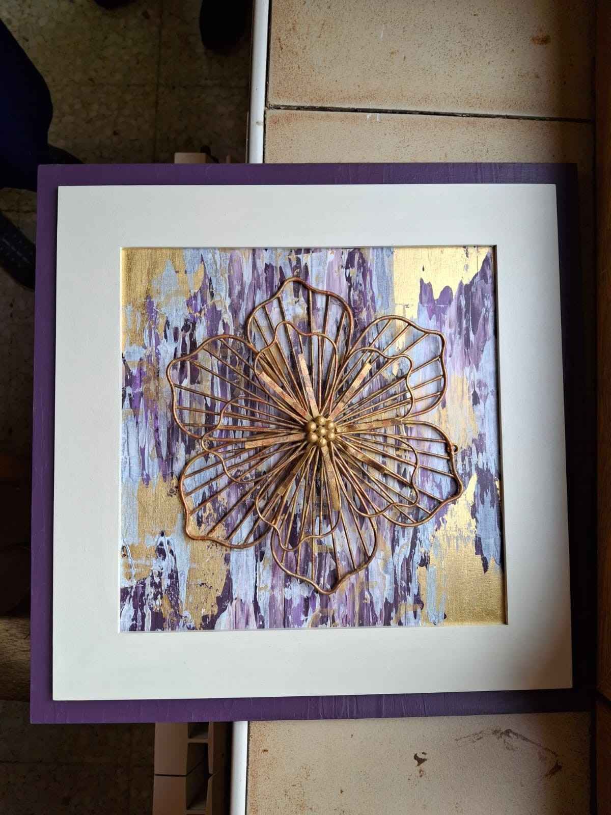 Una obra de arte enmarcada, realizada con técnica mixta, con una flor de alambre dorado metálico sobre un fondo texturizado.