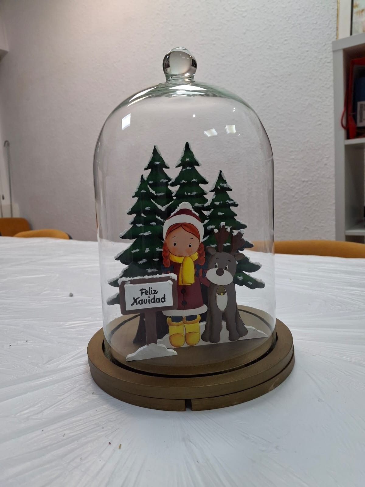 Una campana de cristal muestra una escena festiva con pinos, un personaje con una bufanda amarilla.