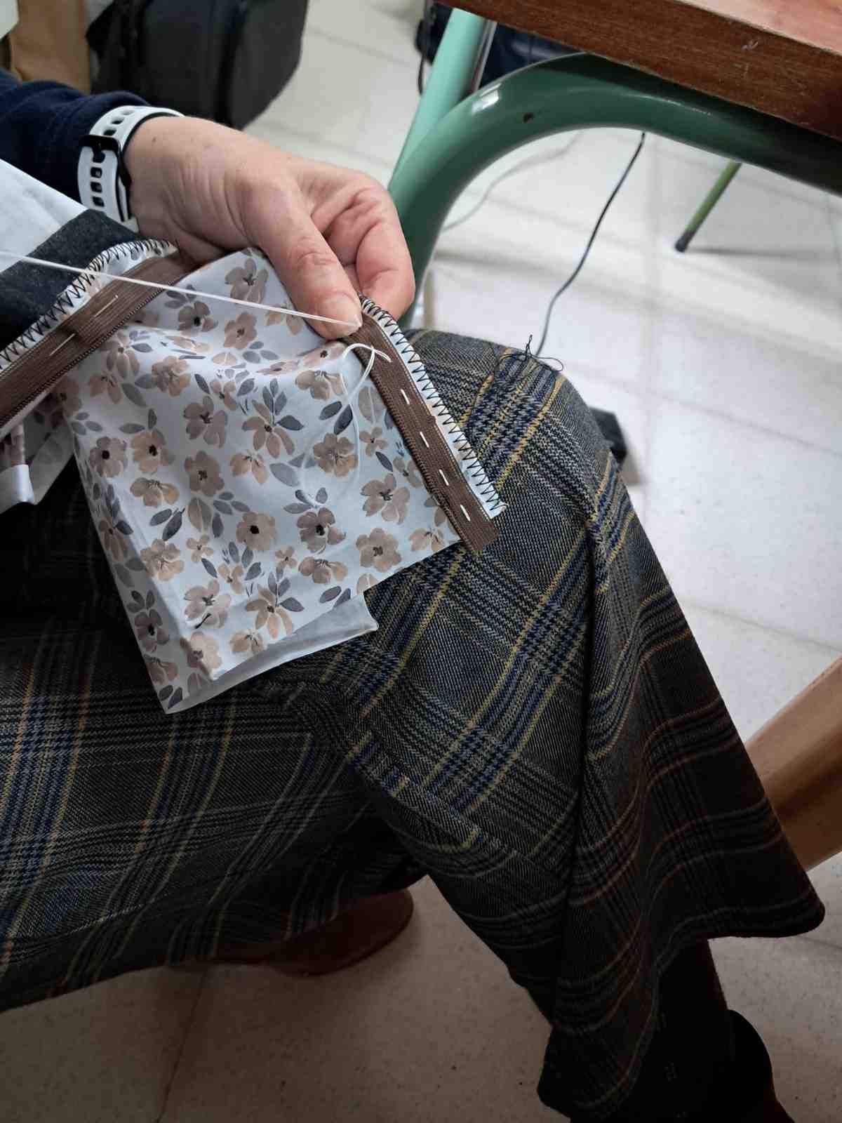 Unas manos cosen tela floral a un borde marrón similar al cuero mientras están sentadas sobre unos pantalones a cuadros.